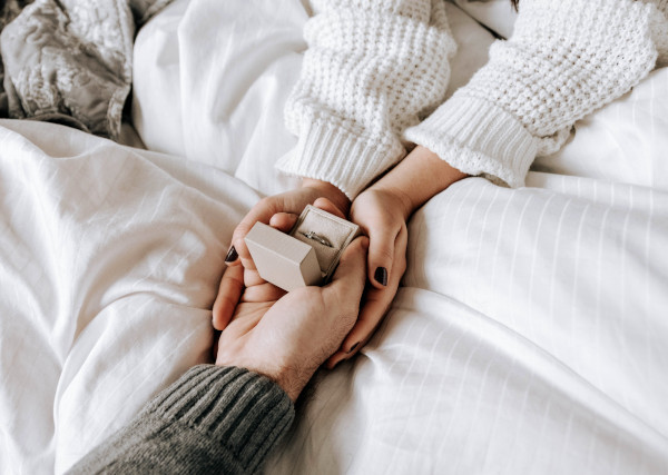 One hand is holding an open box with a ring, and a second pair of hands is holding that hand